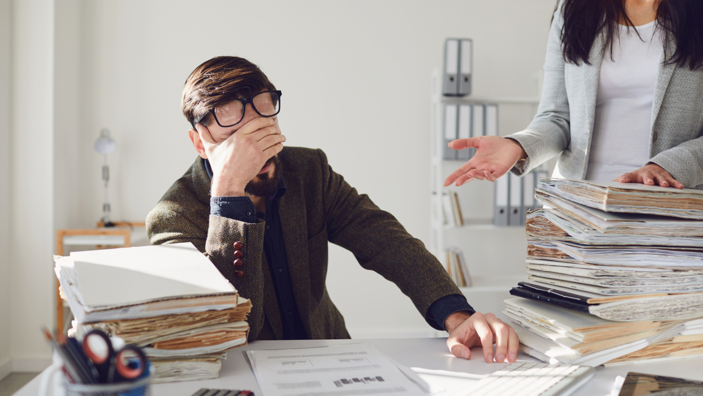 Worker businessman tired upset indignant unhappy sitting at workplace looks at a large amount of work in the office.