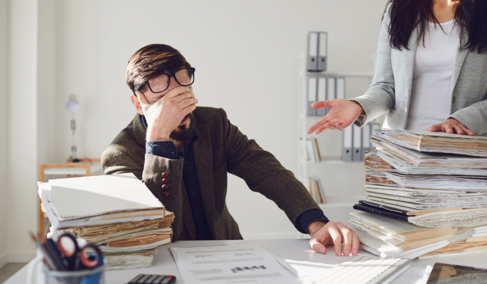 Worker businessman tired upset indignant unhappy sitting at workplace looks at a large amount of work in the office.